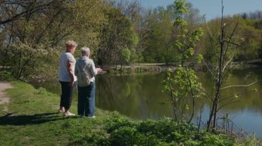 Güneşli bir bahar gününde sakin bir göletin kenarında duran iki yaşlı kadın huzurlu doğanın tadını çıkarıyor ve sessizce sohbet ediyorlar. Yüksek kalite 4k görüntü