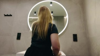 Şık fayanslı bir banyoda yuvarlak bir LED aynayı parlatan sarışın kadın. Yüksek kalite 4k görüntü