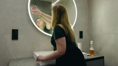 Sarışın bir kadın, temiz, modern bir banyoda parlak, yuvarlak bir LED aynayı siliyor. Yüksek kalite 4k görüntü