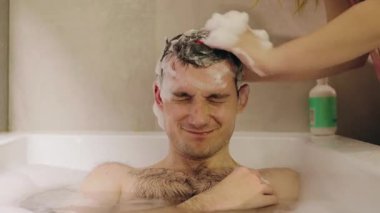 Bir kadın sıcak mum ışığı altında saçlarını köpükle köpüklendirirken mutlu genç adam köpük banyosunda uzanıyor. Yüksek kalite 4k görüntü