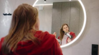 Genç kız saçını kurutuyor ve banyo aynasında yansımasını kontrol ediyor. Yüksek kalite 4k görüntü