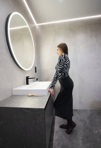 Teenage girl standing in a modern bathroom, leaning on the sink and looking at her reflection in a round illuminated mirror. High quality photo