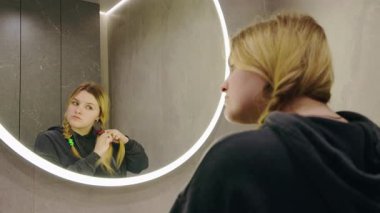 Genç kız şık banyoda parlak yuvarlak aynanın önünde saçını dikkatlice örüyor. Yüksek kalite 4k görüntü