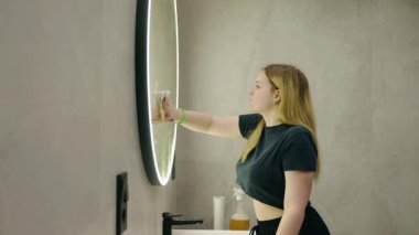 Sarışın bir kadın, temiz, modern bir banyo içinde yuvarlak bir LED aynayı siliyor. Yüksek kalite 4k görüntü