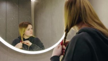 Aydınlanmış banyo aynasında kendine bakarken saçını ören genç bir kız. Yüksek kalite 4k görüntü