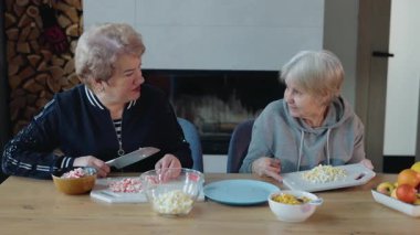 İki yaşlı kadın, sıcak bir mutfakta, ahşap bir masada ev yapımı bir yemek hazırlıyor, sohbet ediyorlar. Yüksek kalite 4k görüntü
