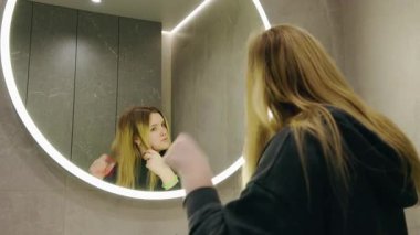 Genç kız modern banyoda aynaya bakarken saçlarını düzeltiyor. Yüksek kalite 4k görüntü