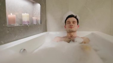 Genç bir adam kafasında köpük bir şapkayla köpük banyosundan zevk alıyor. Rahat ve rahat bir banyo ortamında. Yüksek kalite 4k görüntü