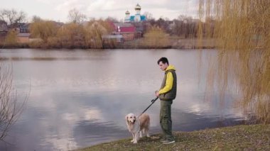Kırsal bir köyde arka planda bir kilise olan gölde Golden Retriever 'ıyla yürüyen genç bir adam. Yüksek kalite 4k görüntü