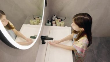 Genç kız, aydınlık bir aynada yansımasına bakarken banyoda ellerini yıkıyor. Yüksek kalite 4k görüntü