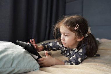 Odaklanmış küçük kız yatağa uzanıyor ve tablet ekranla etkileşime geçiyor, yüzü ciddi ve niyetlidir. Yüksek kalite fotoğraf