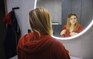 Kırmızı bornozlu genç bir kadın lavaboda dikiliyor, saçına dokunuyor ve aynadaki yansımasını inceliyor. Yüksek kalite fotoğraf