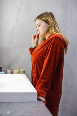 Kırmızı bornozlu genç bir kadın, parlak, modern bir banyoda günlük hijyen rutinini takip ederek, aynanın karşısında dururken dişlerini fırçalar. Yüksek kalite fotoğraf