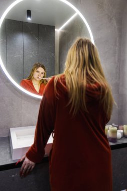Kırmızı bornozlu genç bir kadın şık bir banyoda dişlerini fırçalıyor. Günlük rutini sırasında lavabonun üstündeki parlak aynaya bakıyor. Yüksek kalite fotoğraf