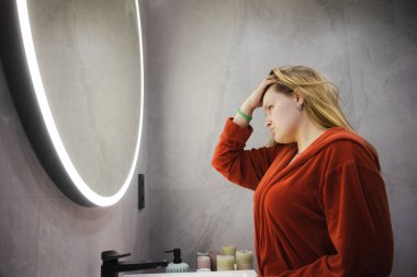Kırmızı bornozlu genç bir kadın elinde cımbızla alnını inceliyor, yumuşak ışıklı modern banyo aynasında yansımasına konsantre oluyor. Yüksek kalite fotoğraf