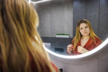 Kırmızı bornozlu genç bir kadın modern bir banyoda diş fırçasını tutarken yuvarlak bir aynada yansımasına dikkatlice bakar. Yüksek kalite fotoğraf