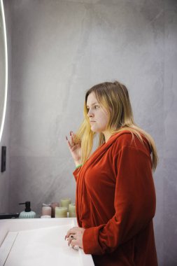 Kırmızı bornozlu genç bir kadın modern bir banyoda lavabonun yanında duruyor. Aydınlatılmış yuvarlak bir aynaya doğru dikkatlice saçına dokunuyor. Yüksek kalite fotoğraf