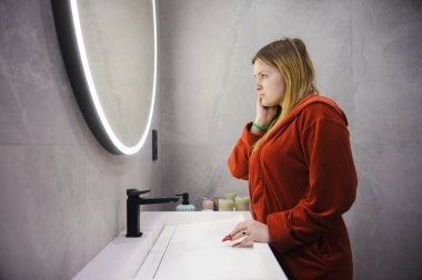 Kırmızı bornozlu genç bir kadın modern bir banyoda, yuvarlak bir LED aynasının önünde yorgun ve düşünceli bir bakışla yansımasına bakıyor. Yüksek kalite fotoğraf