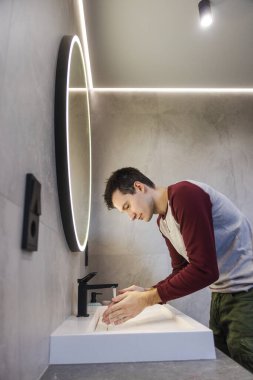 Genç adam ellerini sakince lavaboda yıkıyor. Parlak ve modern bir banyoda temiz ve sağlıklı alışkanlıklarını koruyor. Yüksek kalite fotoğraf