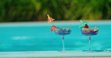 Havuzun kenarında meyve ve şemsiyeli iki renkli yaz kokteyli, turkuaz su arka planına ve yeşil yapraklara karşı. Yüksek kalite 4k görüntü