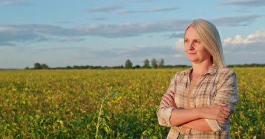 Gün batımında soya tarlasında kolları çapraz duran kendine güvenen dişi çiftçi sakin bir ifadeyle mesafeye bakıyor. Yüksek kalite 4k görüntü