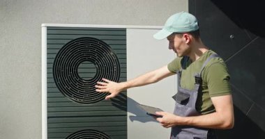 Genç bir erkek teknisyen, elinde tabletle modern bir ısı pompası ünitesinin vantilatörünü inceliyor. Yüksek kalite 4k görüntü