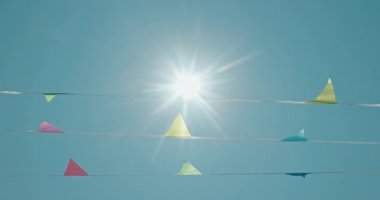 Parlak güneş gökyüzünde asılı renkli üçgen bayraklar boyunca parlayarak yaz kutlamalarının canlı ve neşeli bir atmosferini yaratır. Yüksek kalite 4k görüntü