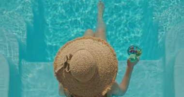 Hasır şapkalı genç bir kadın berrak mavi bir havuzda uzanmış, bacakları açılıp, elinde kokteyl ile huzurlu bir yaz anının tadını çıkarıyor. Yüksek kalite 4k görüntü