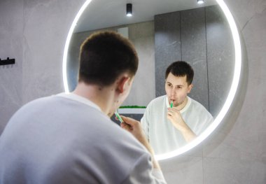 Adam dişlerini parlak yuvarlak bir aynanın önünde fırçalıyor, modern bir banyoda sabah hijyen rutinine odaklanıyor. Yüksek kalite fotoğraf