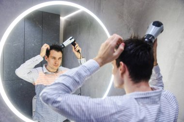 Adam parlak, modern bir banyoda geniş yuvarlak bir LED ışıklı aynanın önünde saçını şekillendirirken saç kurutma makinesi kullanıyor. Yüksek kalite fotoğraf