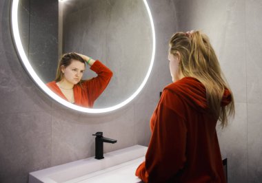 Kırmızı bornozlu genç bir kadın hayal kırıklığına uğramış bir ifadeyle aynada kendine bakıyor, modern bir banyoda LED ışıklandırmasıyla saçlarını topluyor. Yüksek kalite fotoğraf