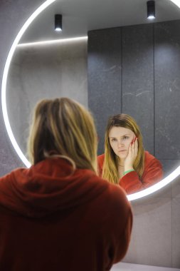 Kırmızı bornozlu genç kadın lavaboya dayanıyor, yüzünü eline yaslıyor ve modern bir banyodaki yuvarlak ışıklı aynaya dikkatlice bakıyor. Yüksek kalite fotoğraf