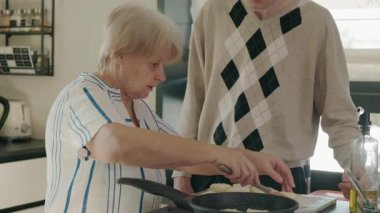 Yaşlı kadın, yetişkin torunu mutfakta yardım ederken süzme peynirli krepleri tavada kızartıyor. Yüksek kalite 4k görüntü