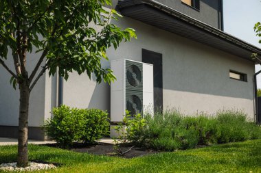 Çevresi yeşil çimenler, çalılar ve bahçedeki genç ağaçlarla çevrili bir ev duvarının yanına kurulmuş modern ısı pompası ünitesi. Yüksek kalite fotoğraf