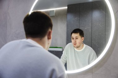 Uykulu ve şaşırmış genç adam, bir sabah rutini sırasında modern bir banyoda duran, yuvarlak ışıklı bir aynada yansımasına bakar. Yüksek kalite fotoğraf