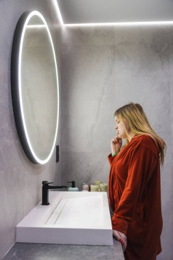 Kırmızı bornozlu genç bir kadın modern bir banyoda parıldayan yuvarlak bir aynada dişlerini fırçalarken lavaboda duruyor. Yüksek kalite fotoğraf