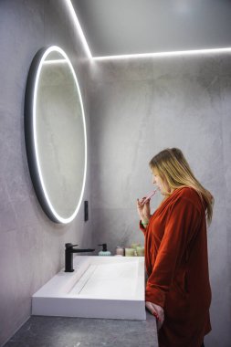 Kırmızı bornozlu genç bir kadın, parlak dairesel bir aynanın önünde dişlerini fırçalıyor, şık, gri renkli bir banyoda yansımasına odaklanıyor. Yüksek kalite fotoğraf
