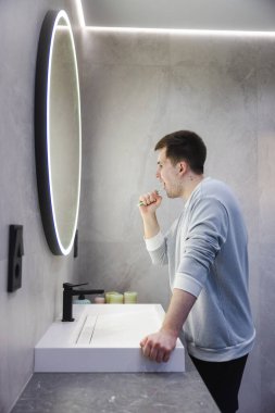 Hafif kazaklı bir adam modern bir banyo lavabosunda dikilirken dişlerini fırçalar, yuvarlak ışıklı aynadaki yansımasına odaklanır. Yüksek kalite fotoğraf