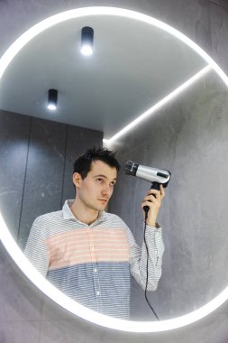 Genç adam parlak yuvarlak bir aynanın önünde duruyor, modern bir banyoda ıslak saçlarını kurutuyor ve şekillendiriyor. Yüksek kalite fotoğraf