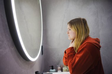 Kırmızı bornozlu genç bir kadın, parlak dairesel bir aynanın önünde yanağını elinde dinlendirirken düşünceli bir ifadeyle yansımasına bakıyor. Yüksek kalite fotoğraf