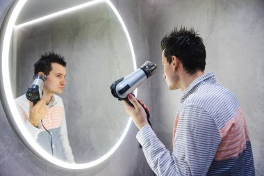 Islak saçlı bir adam, temiz ve modern bir banyo içinde yuvarlak bir LED ışıklı aynaya bakarken saç kurutma makinesi kullanıyor. Yüksek kalite fotoğraf