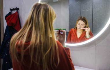 Kırmızı bornozlu genç bir kadın lavaboda durup dişlerini fırçalıyor, parlak, yuvarlak bir aynadaki yansımasına odaklanmış modern bir banyoda. Yüksek kalite fotoğraf