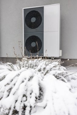 Gri bir duvara monte edilmiş, kışın bahçesinde karla kaplı çalılarla çevrili modern bir açık hava ısı pompasının yakın çekimi. Yüksek kalite fotoğraf