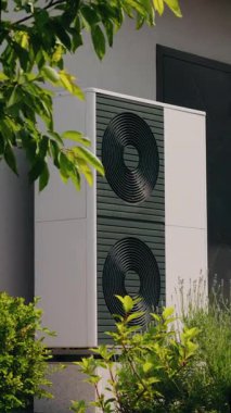 Çevre dostu ev enerjisi çözümlerini vurgulayarak, yeşil yapraklar ve bitkilerle kısmen gizlenmiş, modern bir ısı pompası ünitesinin yakın görüntüsü. Yüksek kalite 4k görüntü