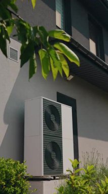Yeşil bitkilerle çevrili ve ağaç yapraklarıyla kısmen gölgelenmiş bir binanın dış duvarına monte edilmiş modern bir ısı pompasının dikey görüntüsü. Yüksek kalite 4k görüntü