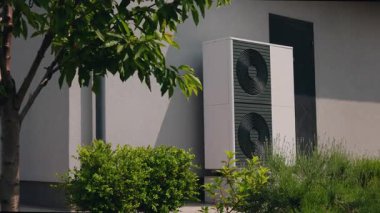 Modern ısı pompası yerleşim yerinin duvarına monte edilmiş, yemyeşil çalılarla çevrili ve peyzaj bahçesindeki bir ağaçla kısmen gölgelenmiş. Yüksek kalite 4k görüntü