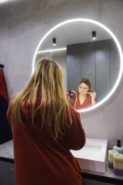 Modern bir banyoda, kırmızı bornozlu genç bir kadın, yuvarlak bir aynanın önünde dişlerini fırçalıyor. Yüksek kalite fotoğraf