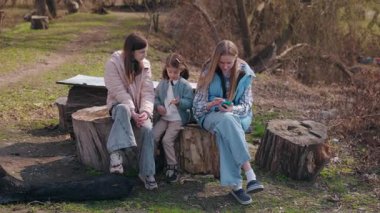 Üç kız, kırsal bir bahar manzarasında ağaç kütüklerinde dinlenirken, bir kız parlak bir şekilde gülümserken, diğerleri bir telefon ve bir yaprağa gömülmüş durumda. Yüksek kalite 4k görüntü