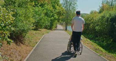 Güneşli bir günde, etrafı yemyeşil olan orta yaşlı bir kadını orman yolunda tekerlekli sandalyeyle iten bir adam. Yüksek kalite 4k görüntü