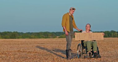 Genç adam tekerlekli sandalyedeki kadını destekliyor. Elinde 